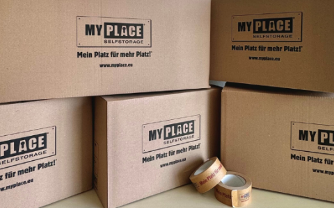 5 verschieden große Kartons sind gestapelt, auf ihnen ist groß das Logo von MyPlace-SelfStorage zu sehen, davor liegen zwei Rollen Klebeband