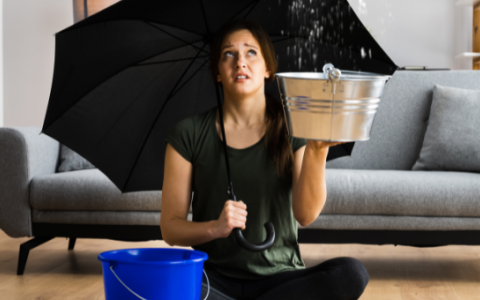 Eine Frau sitzt im Schneidersitz auf dem Boden in ihrem Wohnzimmer, mit einem Regenschirm in der Hand und einem Eimer neben sich stehen. Sie sieht traurig aus und es tropft über ihr von der Decke..