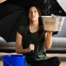 Eine Frau sitzt im Schneidersitz auf dem Boden in ihrem Wohnzimmer, mit einem Regenschirm in der Hand und einem Eimer neben sich stehen. Sie sieht traurig aus und es tropft über ihr von der Decke..